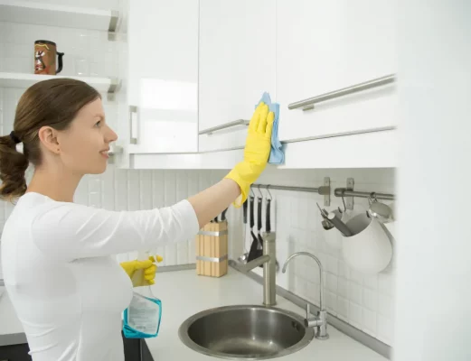 Woman wearing yellow gloves wipes a white kitchen cabinet with a blue cloth; spray bottle on the counter nearby.