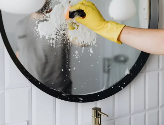 Person wearing yellow gloves wiping a round bathroom mirror with soapy foam visible on the glass.
