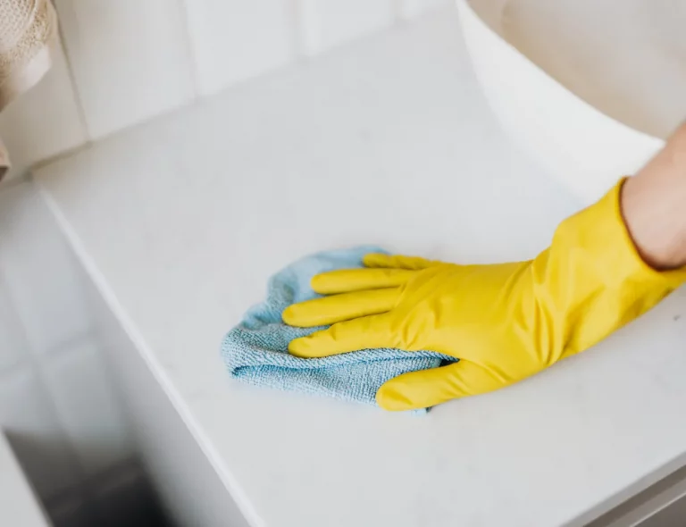 Yellow rubber glove-wearing hand wiping a white countertop with a blue cleaning cloth.