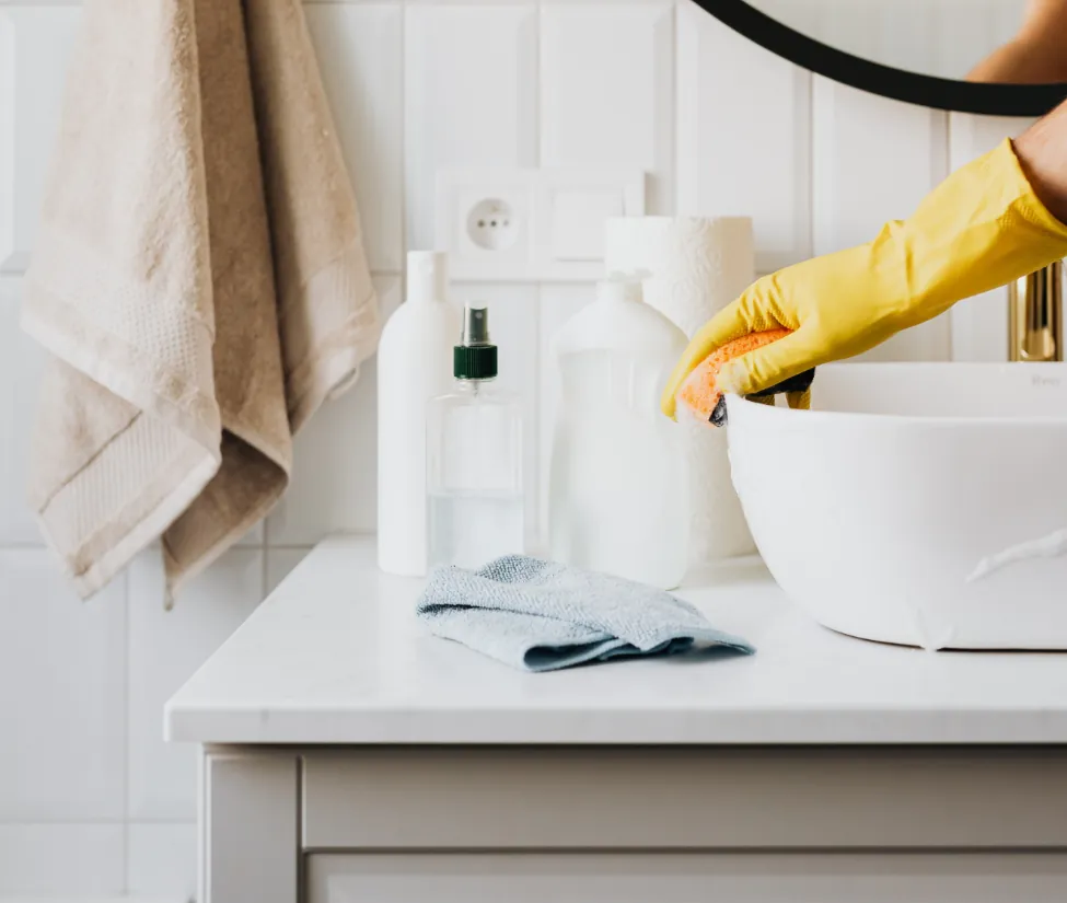 Gloved hand wiping a white bathroom sink with cleaning bottles on the counter nearby.