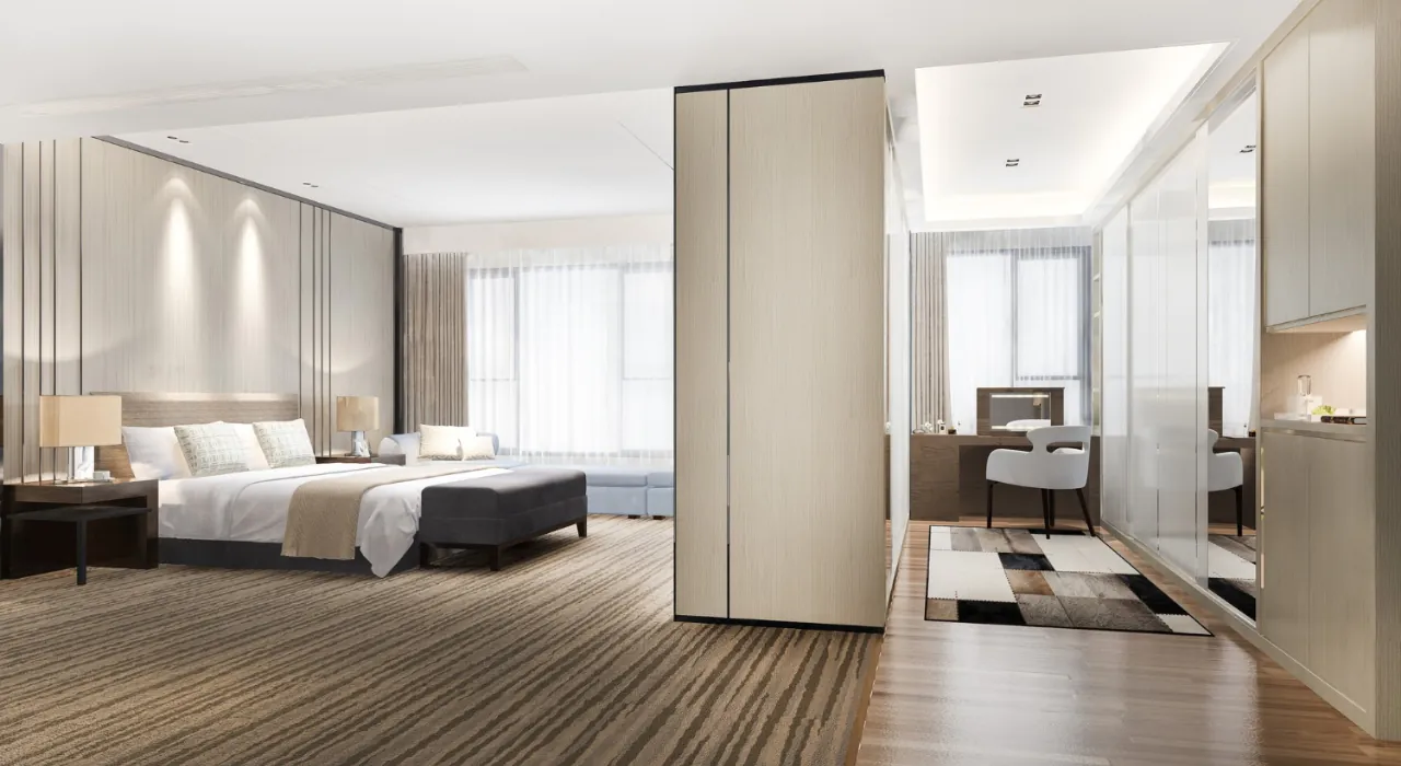 Spacious hotel suite with a king bed, nightstands, and a seating area by large windows, beige decor.