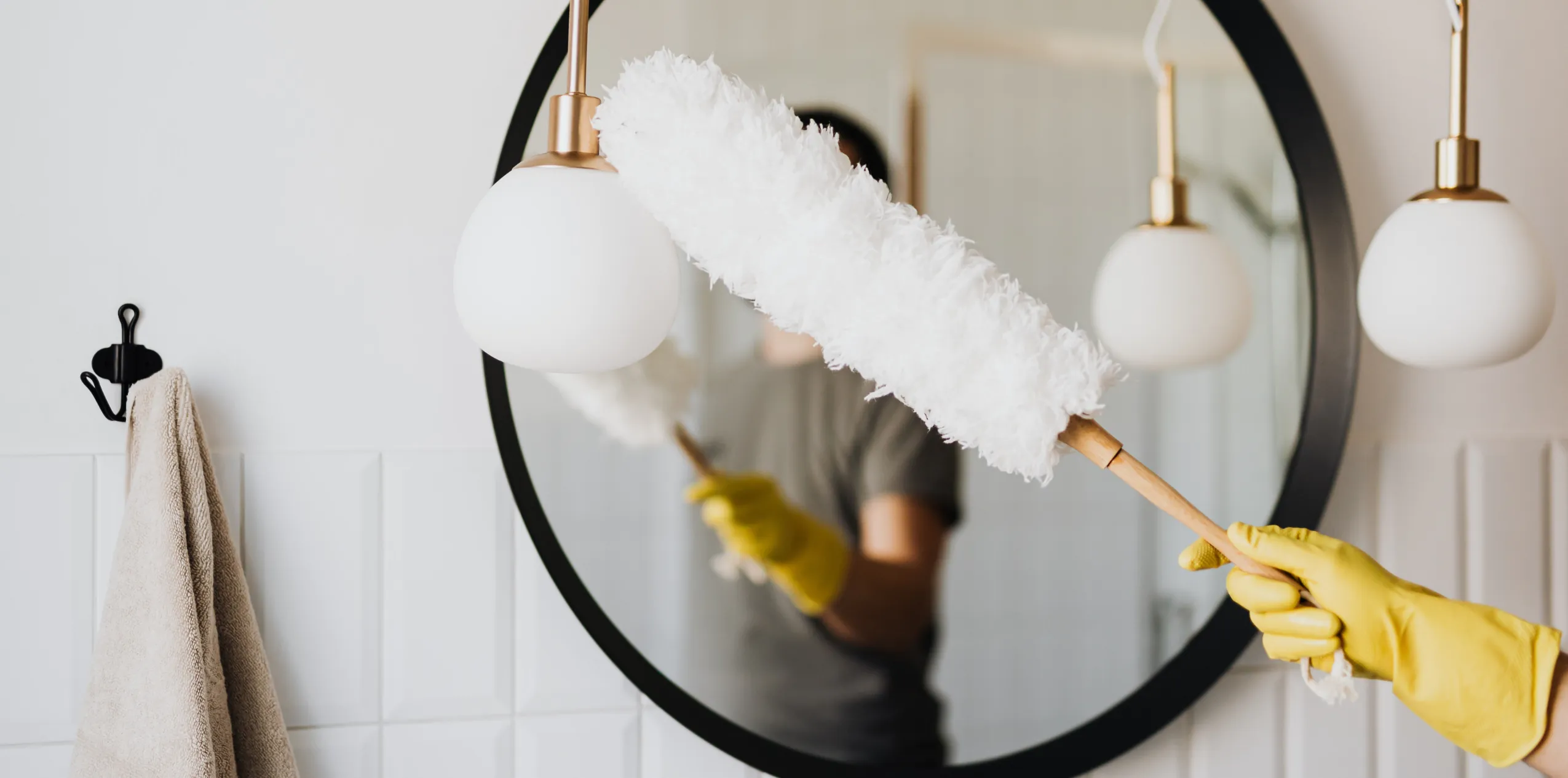 Person wearing yellow gloves dusting a round bathroom mirror with a fluffy white duster in front of it.