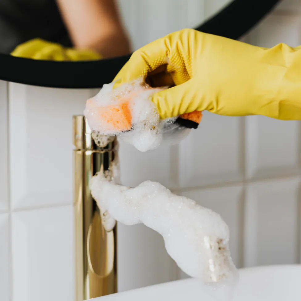 Person wearing yellow gloves scrubbing a gold faucet with soapy foam.