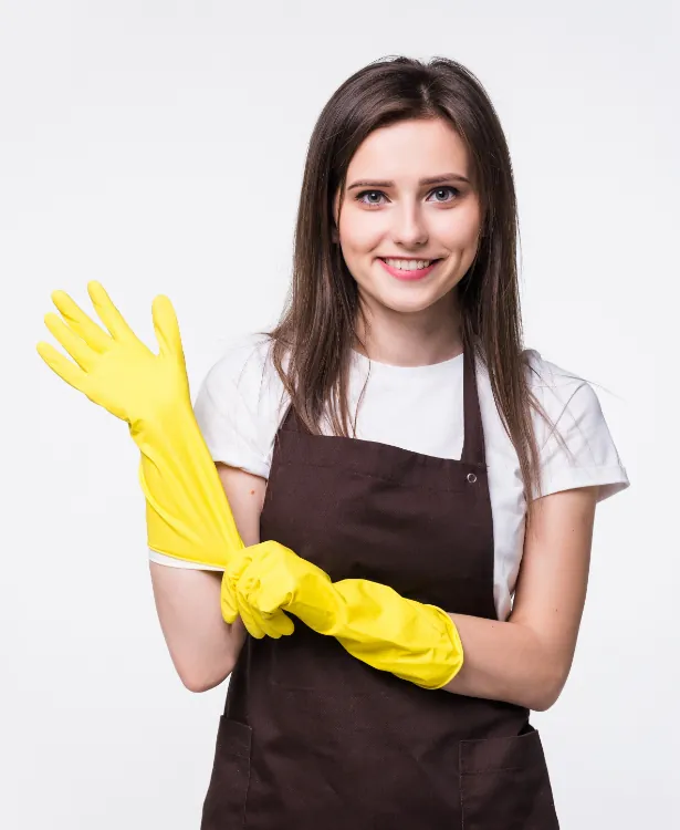 Smiling young woman in a white shirt and brown apron wearing bright yellow rubber gloves, posing with one hand raised.
