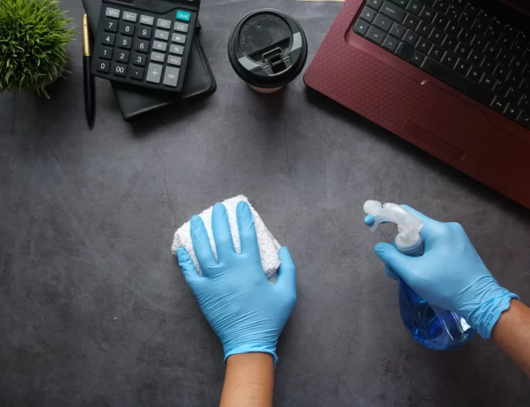 Person in blue gloves wipes a dark desk with a cloth; a spray bottle, laptop, calculator, mug, and plant nearby.