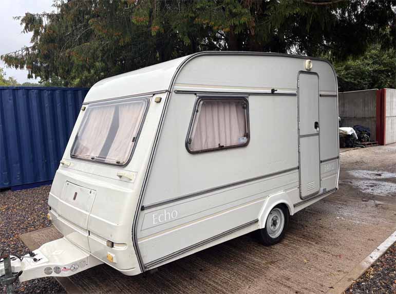 static caravan at yard | Scrap My Caravan White travel trailer (caravan) named Echo, parked on a concrete lot with a tow hitch and pink-curtained windows.
